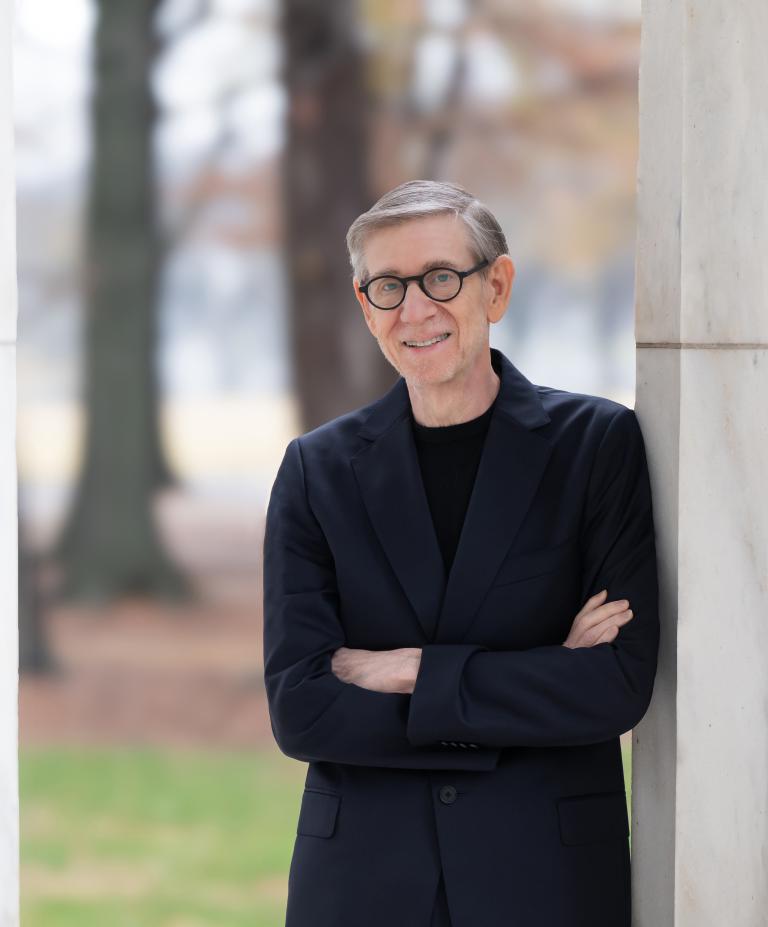 A man in a black shirt and suit jacket with thick rimmed glasses leans against a marble column outside with arms crossed and smiles slightly at the camera