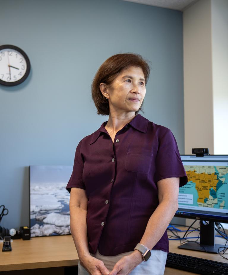 A woman stands in an office in front of two computer screens and looks toward the window with a serious expression.
