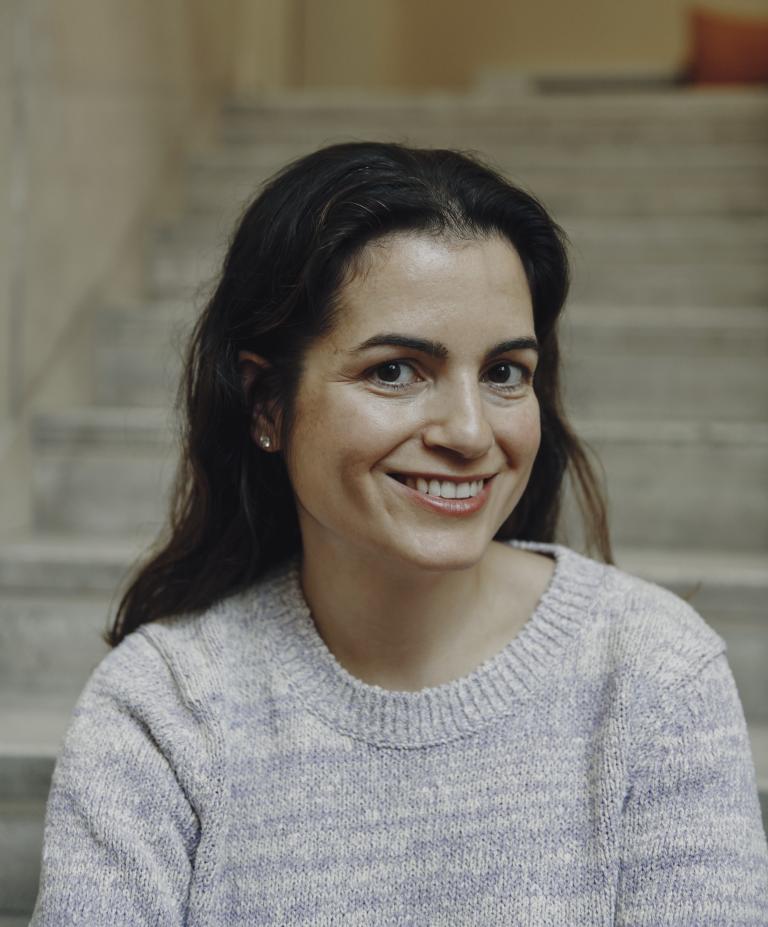 Smiling woman with dark hair wearing a light sweater, sitting on stone steps. The background is softly blurred, conveying a warm and inviting atmosphere.