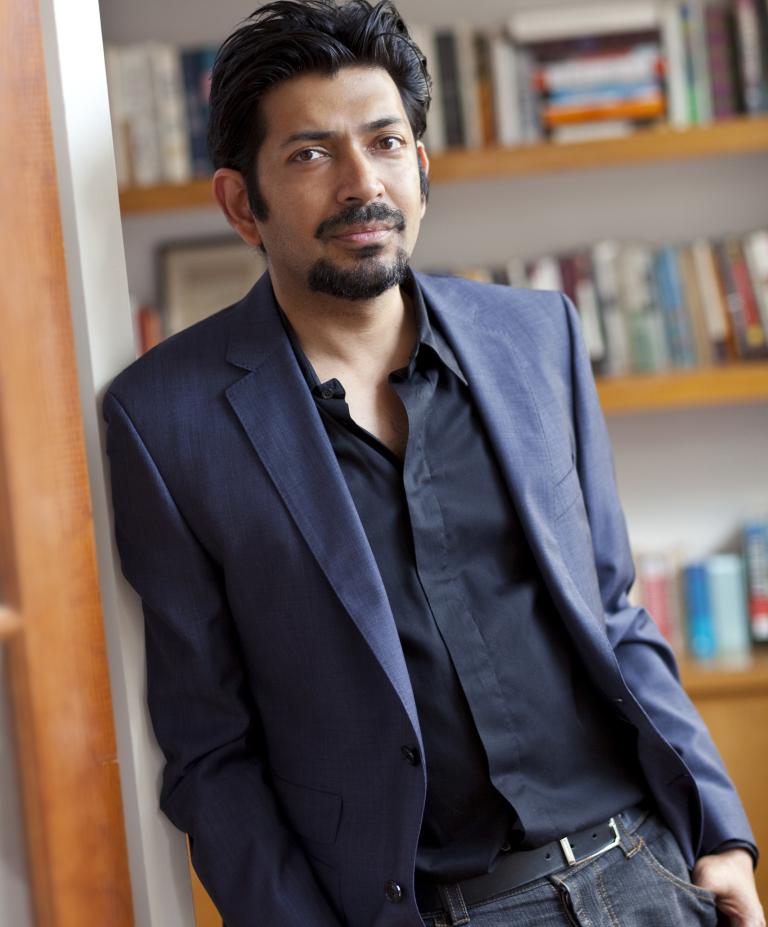 Siddhartha Mukherjee in a navy suit leans against a wall beside a bookshelf.
