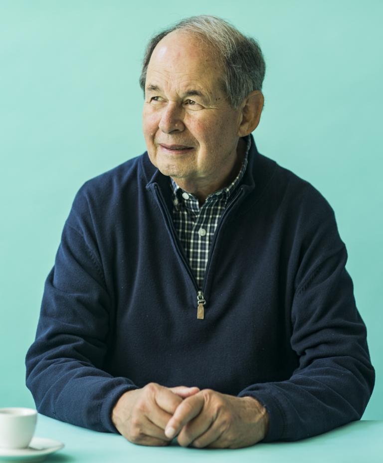 A man in a navy sweater sits at a teal table with a thoughtful expression. A cup of coffee is placed in front of him, against a teal background.