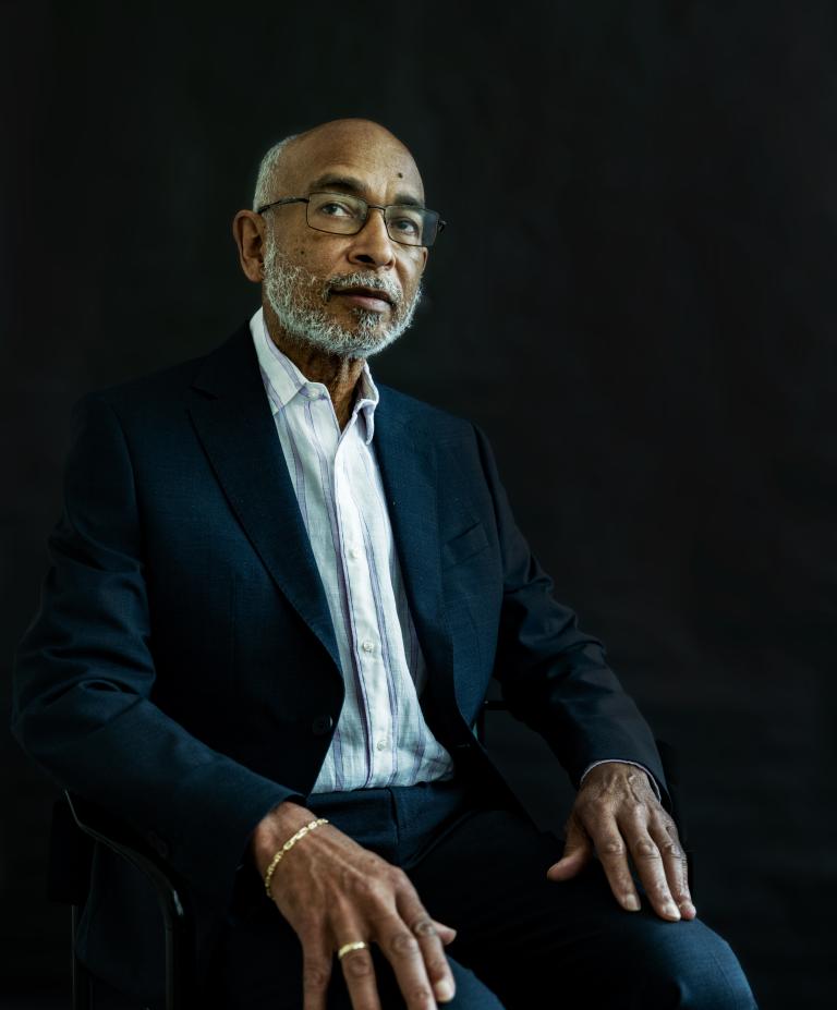 A portrait of an older man in a suit sitting on a chair, looking thoughtfully to the side, against a stark black background.