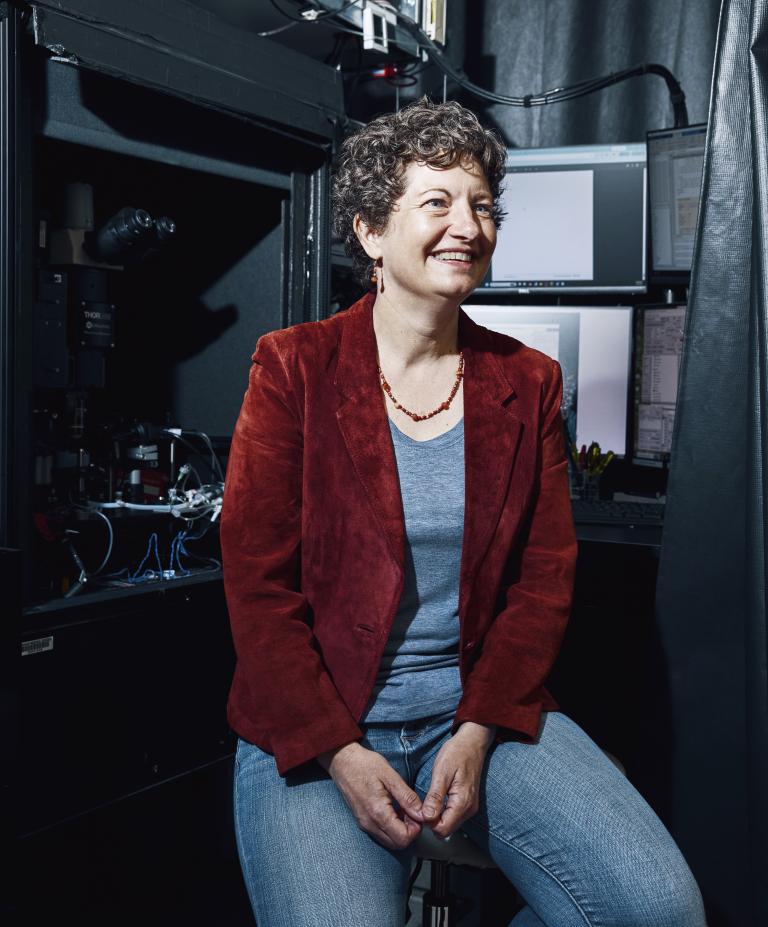 A woman with short curly hair, wearing a red blazer and jeans, sits on a stool in a lab with computers and scientific equipment in the background. She has a wide smile and is looking off to the side.