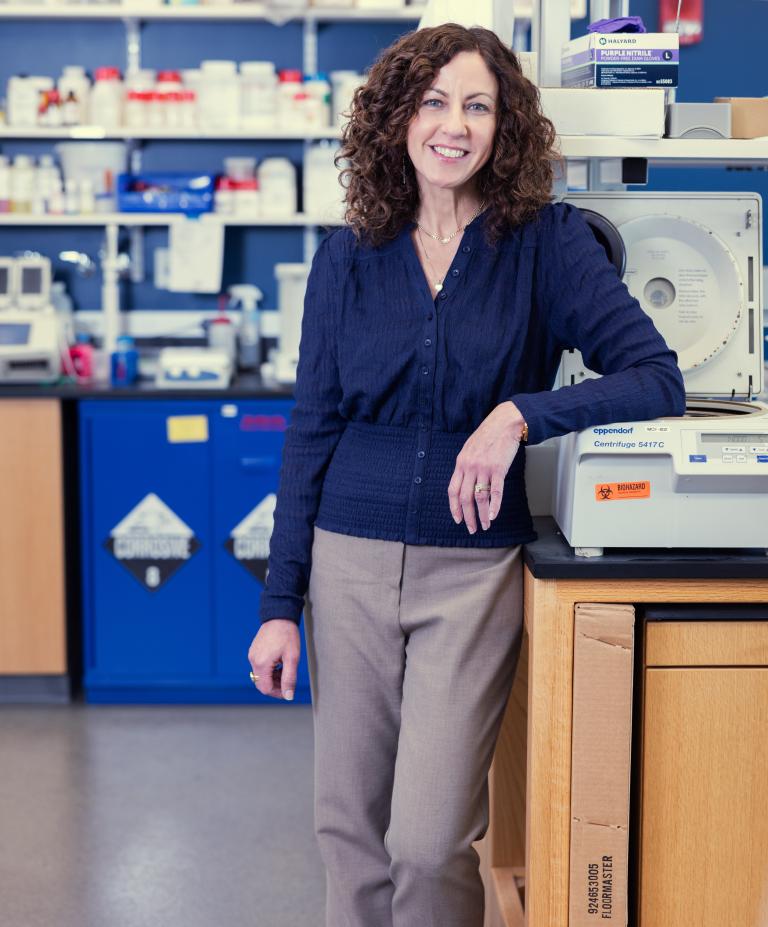 Smiling woman standing a laboratory surrounded by glassware, scientific supplies, and equipment.