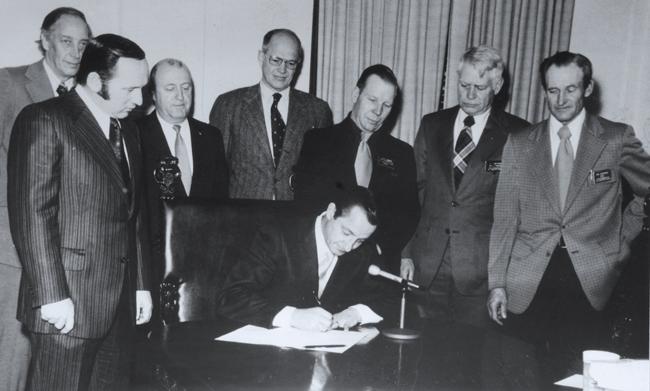 black and white photo of men standing over the governor signing a bill at his desk