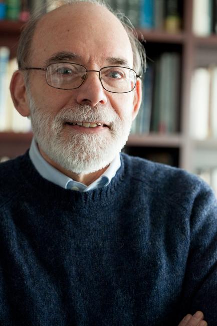 man in blue sweater with glasses and white beard in front of bookshelf