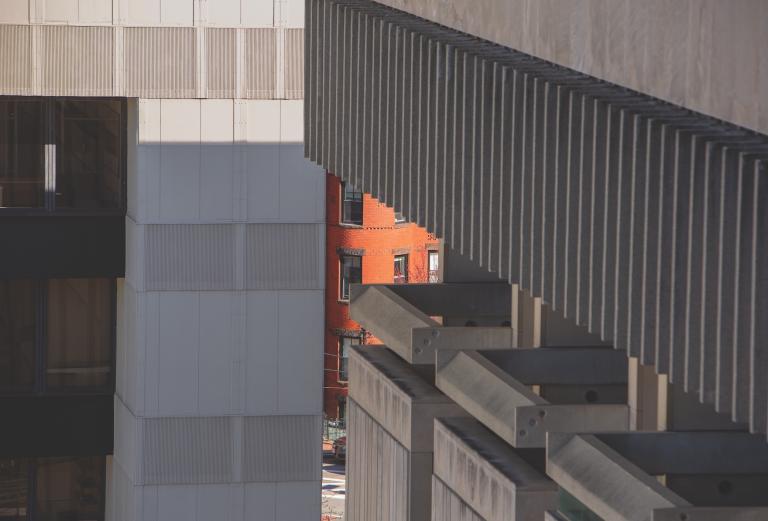 A gap in a concrete, brutalist building in the foreground reveals an older red brick building across the street behind it