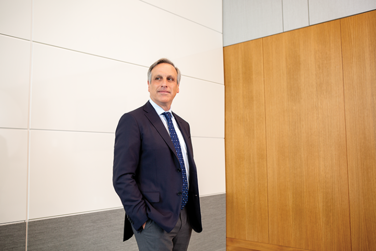 Peter Zimetbaum stands in a dark suit and tie in a room with a wood-paneled wall on the right and white wall on the left
