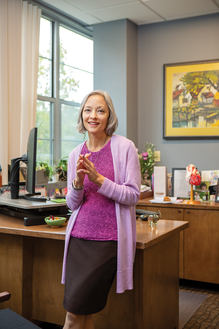 Dara Lee Lewis leans against a wooden desk in an office. She is wearing a pink shirt and light pink sweater, and smiles for the camera.