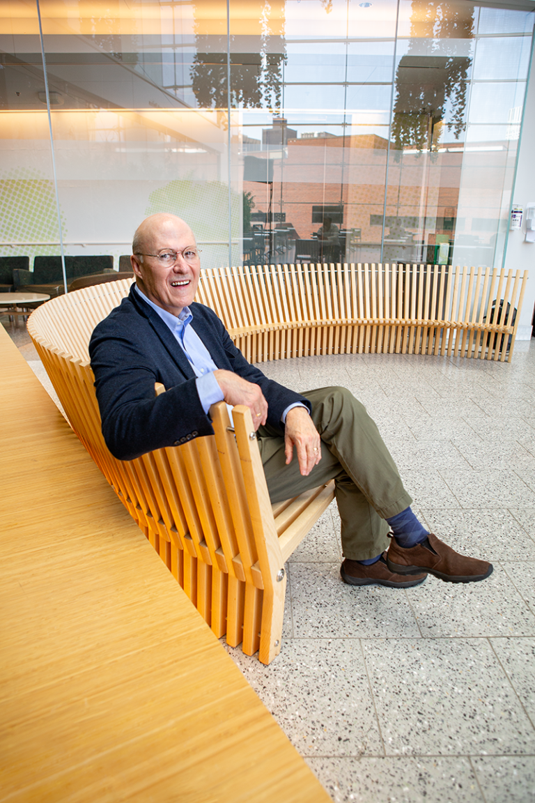 Gregory Fricchione sits on a long, curved wooden bench inside a building with a glass wall, smiling at the camera.