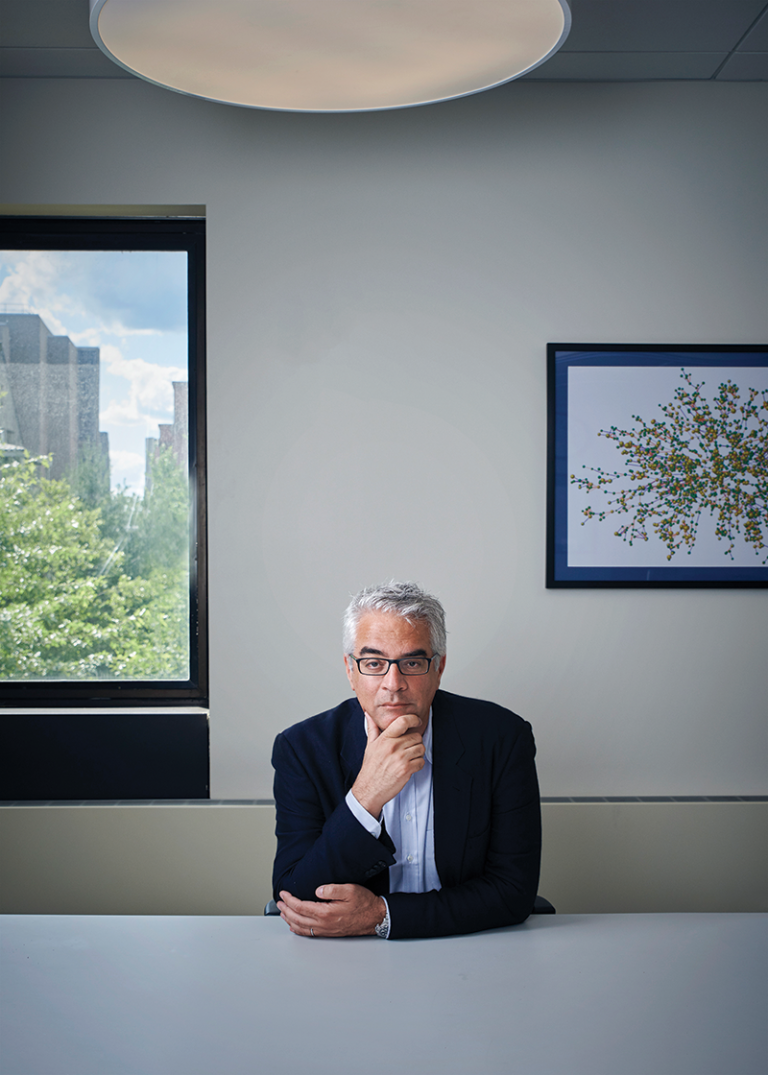 Nicholas Christakis sits at a table in an off-white room between a window looking out onto a city and a framed piece of art. He is looking inquisitively at the camera.