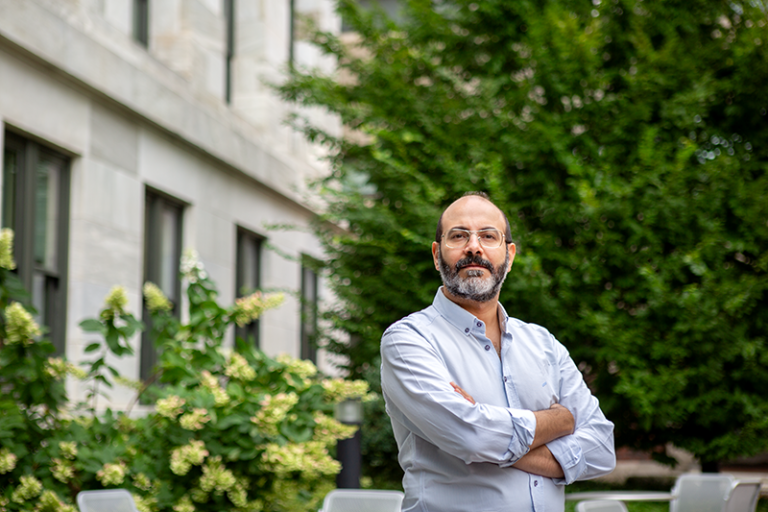 Michael Elnemais Fawzy stands with arms crossed, looking at the camera, outside of a building framed by green trees and bushes.