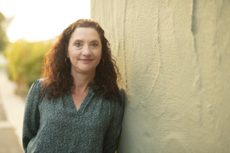 Janine Young, a woman with curly brown hair, looks at the camera while leaning against a beige wall.