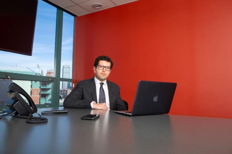 Alex Keuroghlian sits at a table in a room with a red wall and a large window out onto the city. He is wearing a suit jacket and tie and looking at the camera.