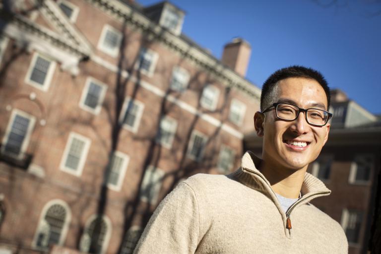 Michael Liu stands in front of a brick building on Harvard campus looking at the camera on a sunny day.