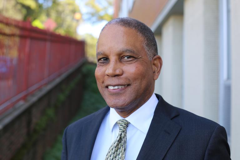 Neil Powe looks at the camera wearing a suit jacket and tie, standing outside of a building next to a red fence