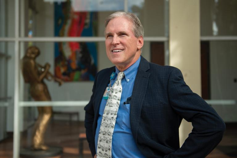 Patrick Whelan smiles wearing a blue shirt and tie with a musical score, in front of a window into a building with a statue and painting.