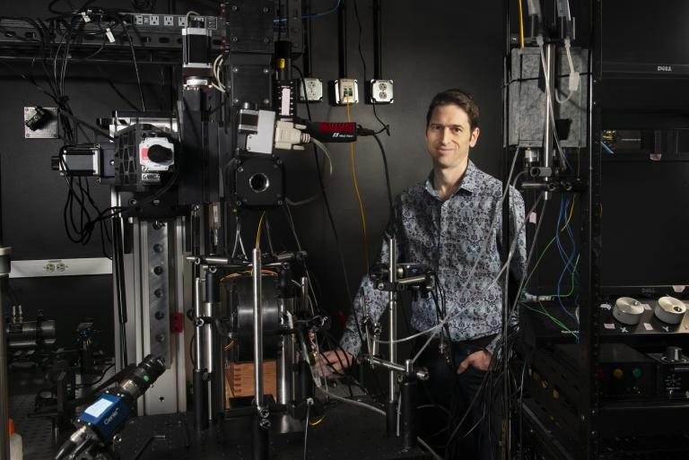 Mark Andermann peeks out from behind a large assortment of laboratory equipment in a dark-colored room.