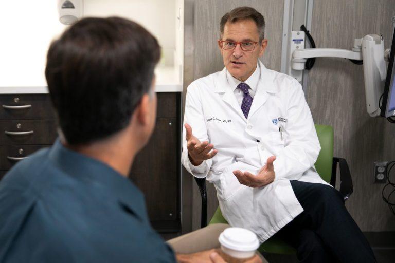 A male doctor in a white coat and tie underneath counsels a patient while sitting down in an office.