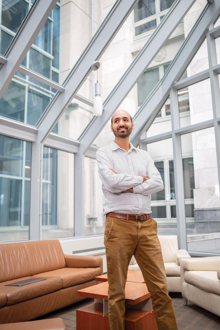 A man in khakis and a white button down shirt stands, arms crossed, looking to the side with a slight smile. He is in a room full of windows.