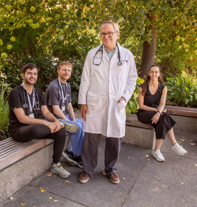 A man in a white coat with a stethoscope stands in front of a bench where three younger people sit. All are smiling at the camera.
