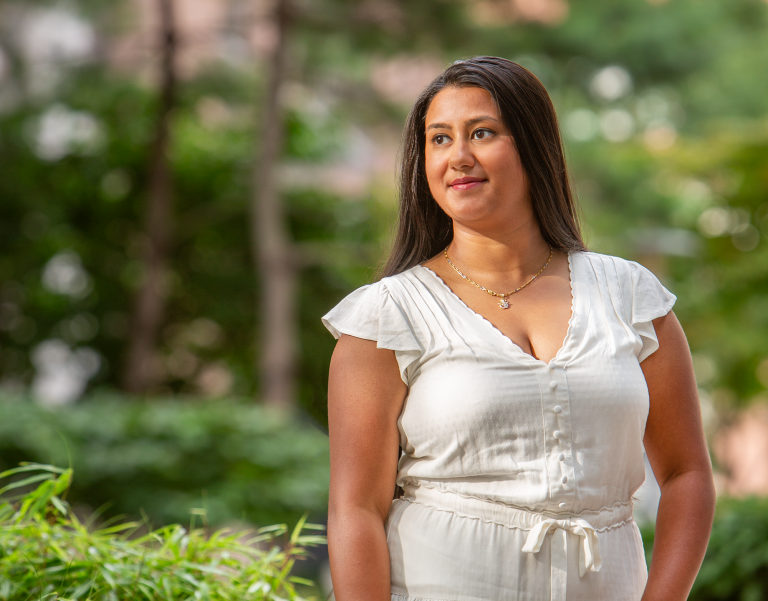 A woman in a white dress stands outside amongst greenery, looking to her right and smiling slightly.