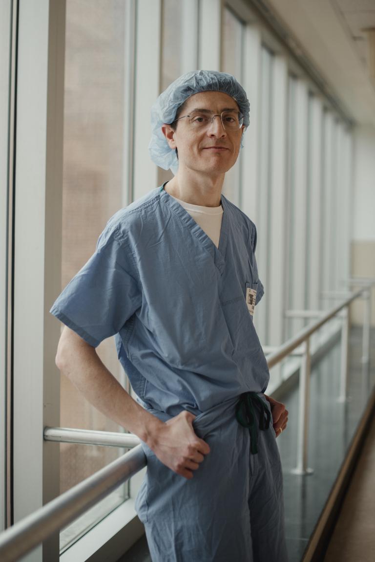 A man in blue scrubs leans against a railing in front of a window, looking at the camera