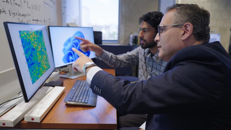 Two men sit at a desk with two monitors, pointing to images on computers