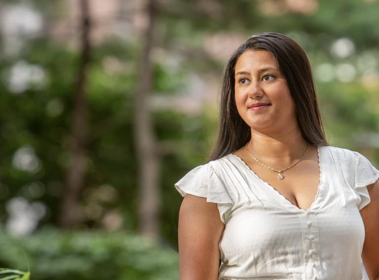 A woman in a white dress stands outside amongst greenery, looking to her right and smiling slightly.