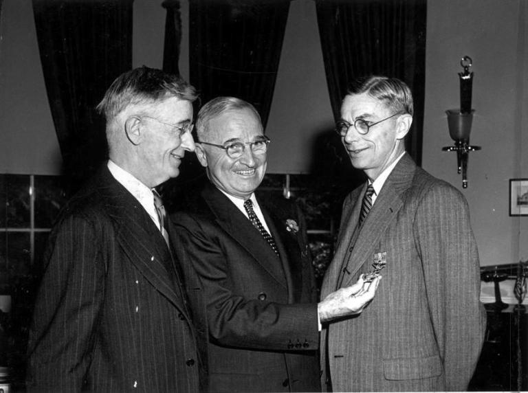 Three men in suits. One of them is examining a medal pinned to another man’s jacket.