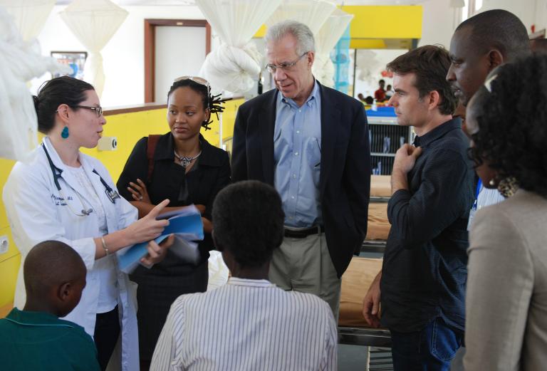A female doctor in a white coat speaks with a group of visitors inside a hospital ward. The group includes a mix of professionals attentively listening, standing near hospital beds.