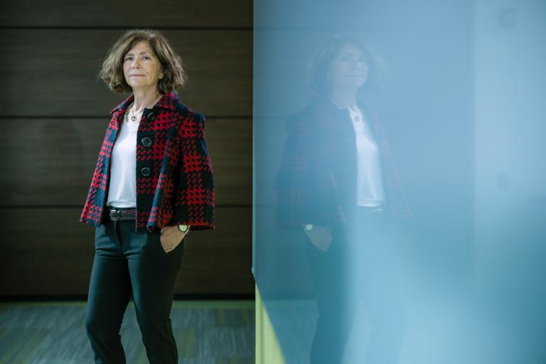 A woman stands confidently in a dark room, wearing a red and black plaid jacket and a white top. She's reflected in a glass panel.