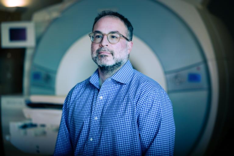A man in glasses and a blue checked shirt stands confidently in front of an MRI machine in a dimly lit room.