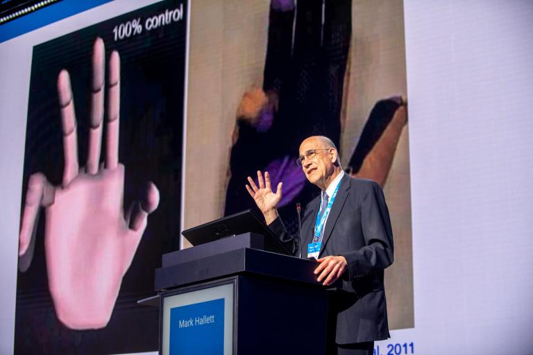 A man in a suit speaks at a conference podium with his hand raised, demonstrating a tremor, as a presentation screen behind him shows a computer-generated image of a hand