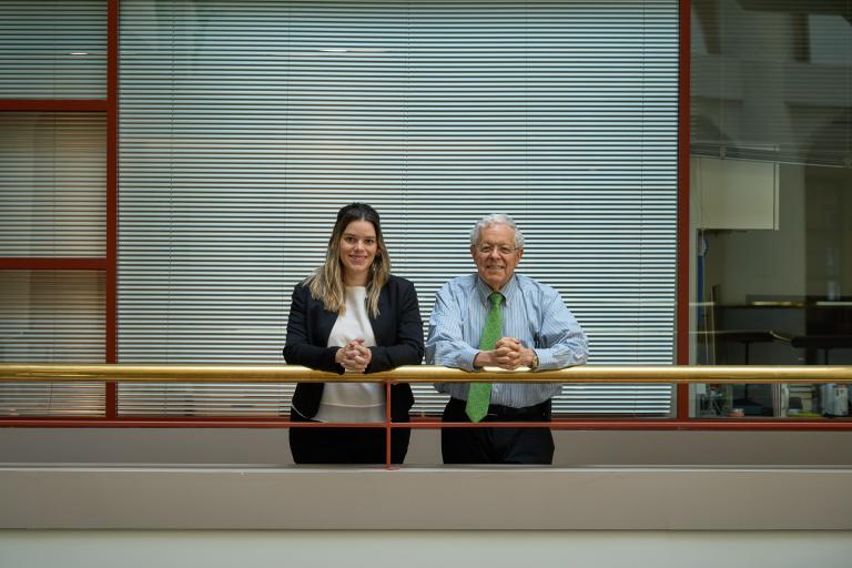 Two people, a woman and an older man, stand side by side smiling with arms on a railing. Background features vertical blinds and a modern office setting.