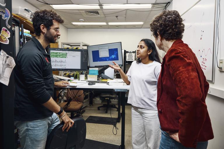 Three people are engaged in a discussion in a laboratory. The background includes a variety of lab equipment, computer monitors, and notes and drawings on a whiteboard.