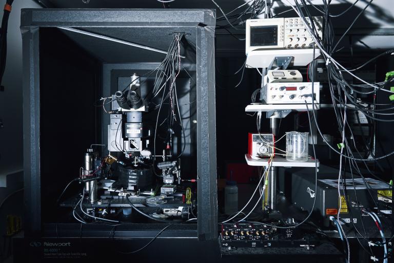 A complex laboratory setup featuring a high-tech microscope enclosed in a black frame structure on the left side. To the right, there is a tall rack with various electronic devices stacked on metal shelves, all connected by a network of tangled cables.