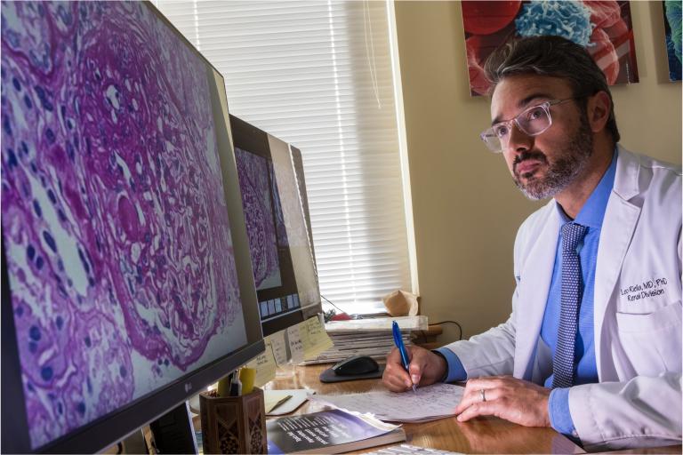 A doctor in a lab coat looks intently at two computer monitors displaying microscopic images, while taking notes at his desk.