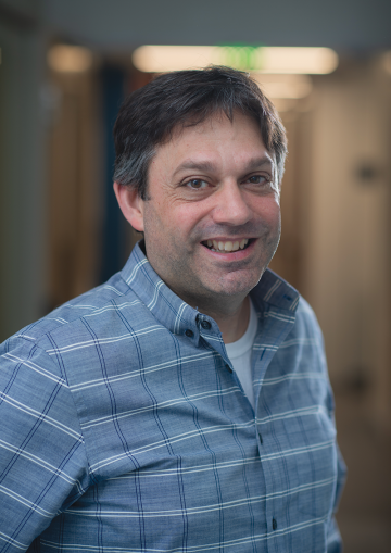 Stephen Liberles stands in a hallway and looks at the camera, smiling, wearing a blue checkered shirt