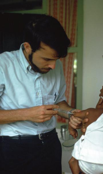 A dark haired young man in a white button down shirt administers a shot to the arm of a baby.