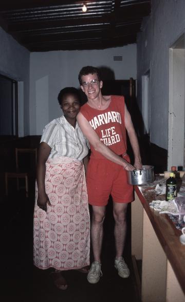 A man in a Harvard Lifeguard shirt stands next to a woman in white shirt and pink skirt in a kitchen. Both are smiling.