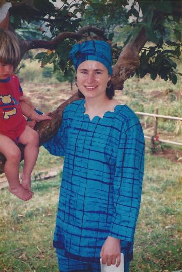 A woman in a blue traditional African outfit stands next to a tree, smiling