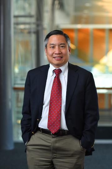 A man in a red tie and black suit jacket looks at the camera smiling with hands in his pockets