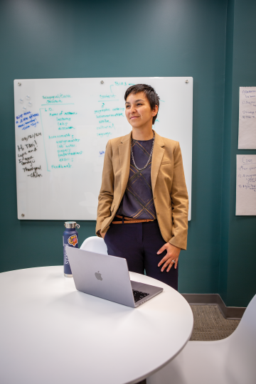 A woman in a beige suit jacket stands in front of a white board behind a table with a laptop on it