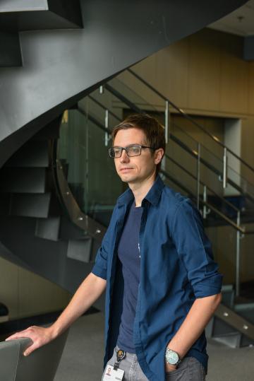 A young man with glasses and a blue button down shirt leans on a railing looking out the window by a winding staircase