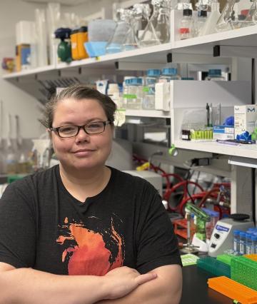 A woman in a black t-shirt with glasses standing in a lab looks at the camera with her arms crossed.