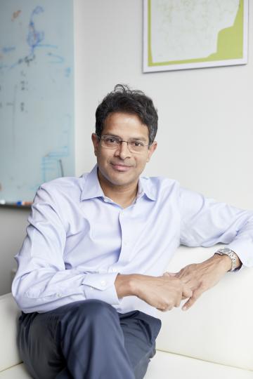 A man wearing glasses and a light blue shirt sits on a white couch with a whiteboard in the background.