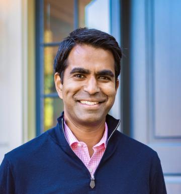 Smiling man in a navy sweater over a pink checkered shirt, standing in front of a blurred blue and beige background. The tone is warm and friendly.