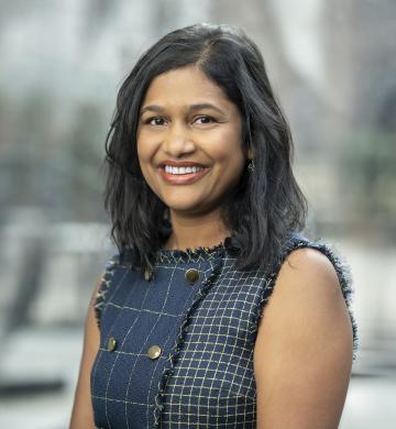 A woman with shoulder-length black hair smiles warmly, wearing a blue patterned sleeveless shirt against a blurred outdoor background