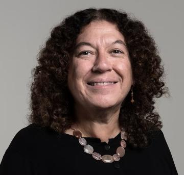 A headshot of a woman, Perri Klass, with brown curly hair, smiling, in a black shirt with a thick gemstone necklace. 